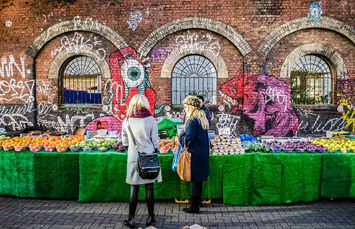 East London'da, Brick Lane'in yanındaki bir meyve tezgahının başında duran iki genç kız.